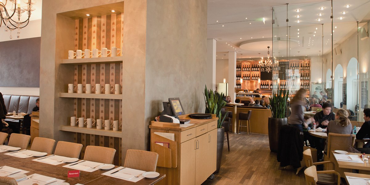 Stylish restaurant with wooden furniture, shelves full of cups and a cozy atmosphere. Guests sit at tables, with a bar in the background., © Aspectus Photographie Stuttgart