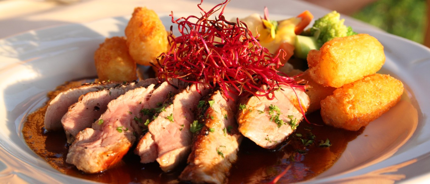 Ein Teller mit saftigem Fleisch, knusprigen Kroketten und Gemüse, garniert mit roten Sprossen, in einem eleganten Restaurant serviert., © Badhotel Restaurant Stauferland