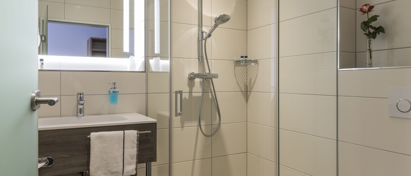 Modernes Badezimmer mit Glasdusche, Waschbecken und Toilette. Helle Fliesen, Spiegel mit Beleuchtung und eine Rose als Dekoration., © Hotel Hirsch