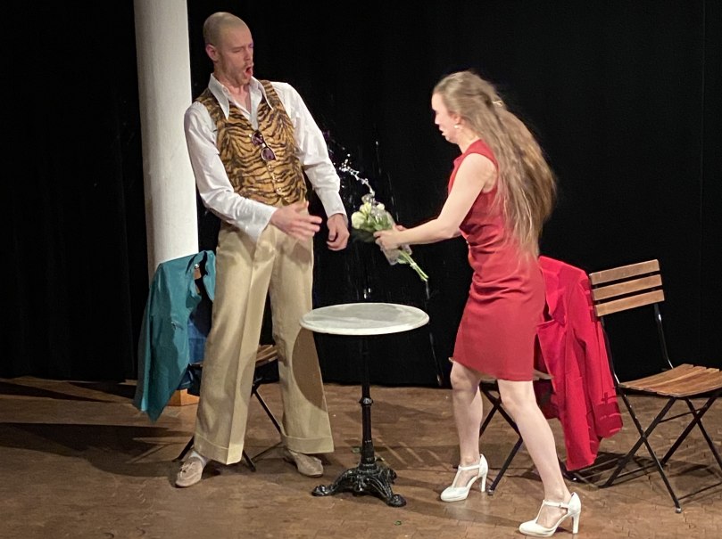Eine Frau in rotem Kleid spritzt Wasser aus einem Blumenstrauß auf einen Mann in Tigerweste auf einer Bühne. Ein Tisch und Stühle sind im Hintergrund., © Wilhelma Theater