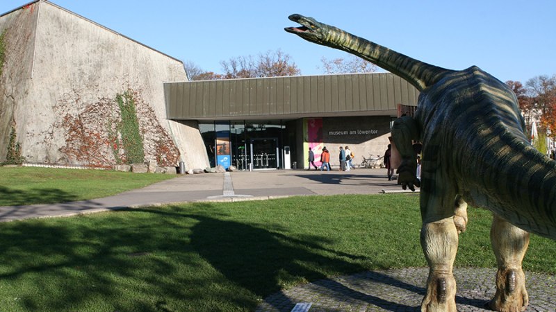 Das Museum am L&ouml;wentor mit einer Dinosaurierstatue im Vordergrund und Besuchern am Eingang., &copy; SMNS, R. Baumann