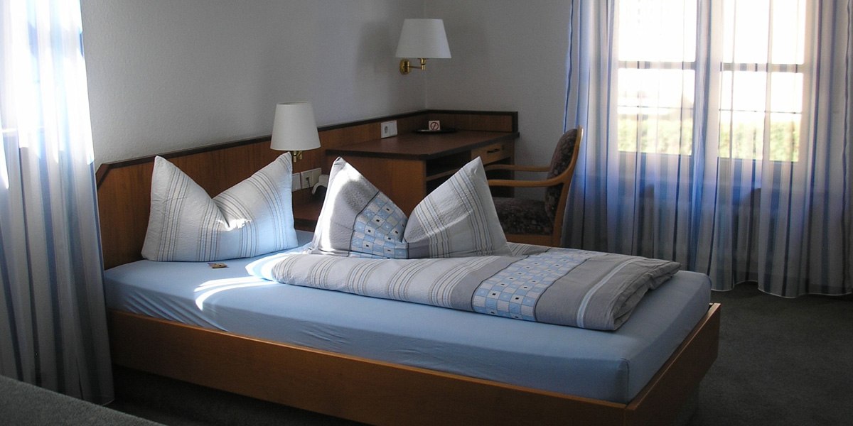 Ein Hotelzimmer mit Einzelbett, blauer Bettwäsche, Schreibtisch und Stuhl. Licht fällt durch gestreifte Vorhänge., © Hotel am Brunnen