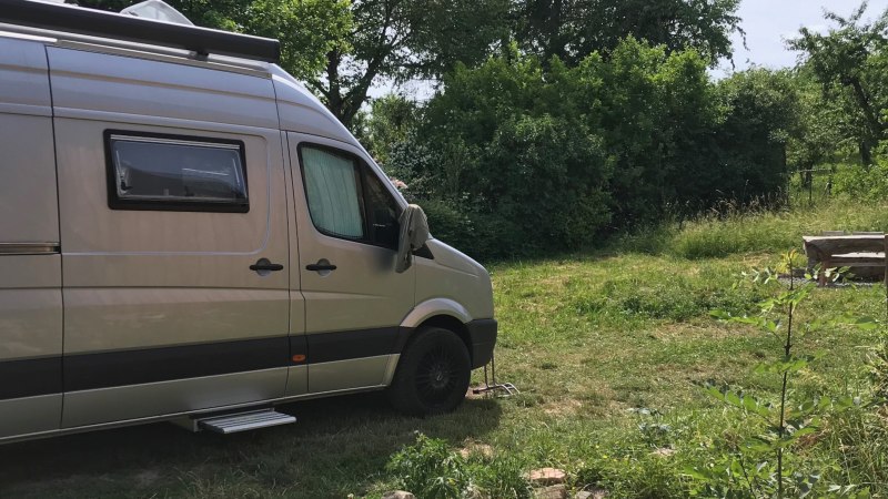 Ein silbernes Wohnmobil steht auf einem grünen Stellplatz. Im Hintergrund sind Bäume und eine rustikale Bank zu sehen., © Weingut Idler