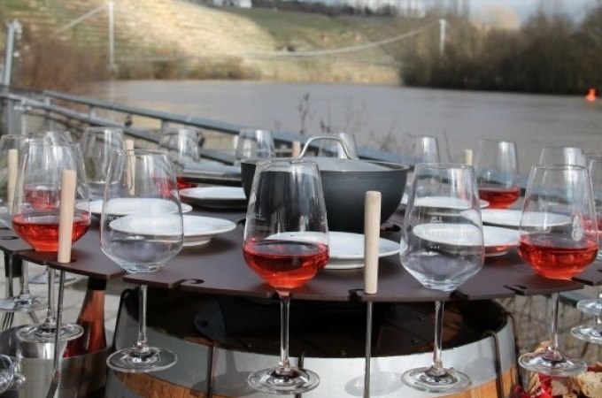 Ein Fass mit Fondue-Set und Weingläsern, gefüllt mit Roséwein, steht im Freien vor einem Fluss und einer Hügellandschaft., © Cool-Tours StattReisen Ein Fass mit Fondue-Set und Weingläsern, gefüllt mit Roséwein, steht im Freien vor einem Fluss und einer Hügellandschaft., © Cool-Tours StattReisen