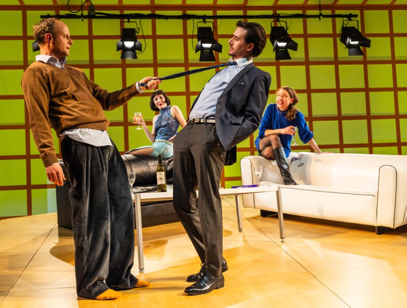 Vier Personen in einem modernen Wohnzimmer-Setting. Zwei M&auml;nner stehen im Vordergrund, einer zieht am Schlips des anderen. Zwei Frauen sitzen entspannt im Hintergrund., &copy; W&uuml;rttembergische Staatstheater Stuttgart