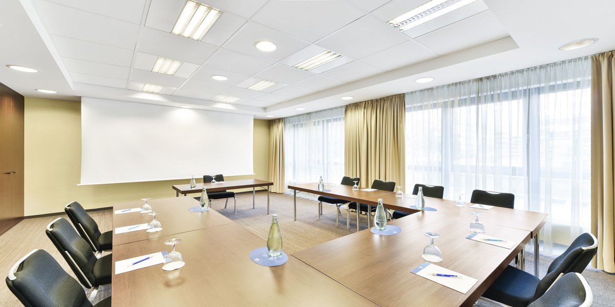 A bright conference room with a U-shaped table arrangement, chairs, water bottles, and writing supplies. Large windows with curtains let in plenty of light., © NH Ludwigsburg A bright conference room with a U-shaped table arrangement, chairs, water bottles, and writing supplies. Large windows with curtains let in plenty of light., © NH Ludwigsburg