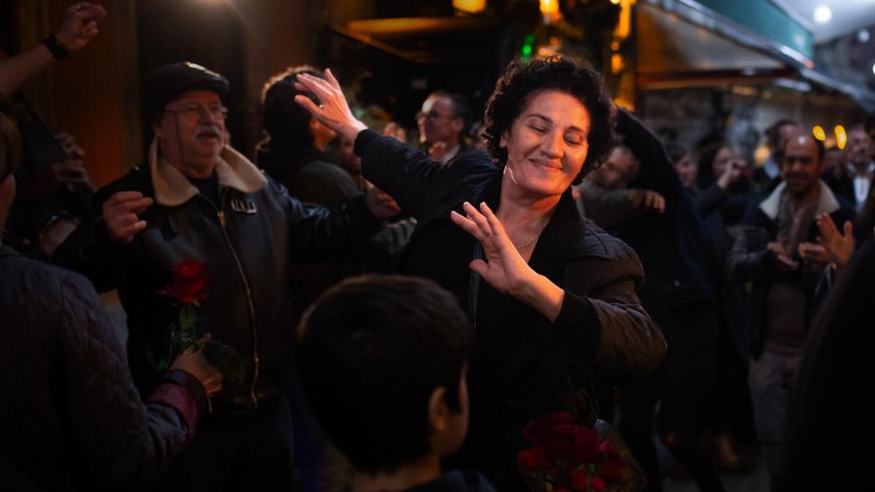 Eine Frau tanzt lächelnd in einer Menschenmenge auf einer belebten Straße bei Nacht. Andere Personen halten Rosen und feiern mit., © MUBI Eine Frau tanzt lächelnd in einer Menschenmenge auf einer belebten Straße bei Nacht. Andere Personen halten Rosen und feiern mit., © MUBI