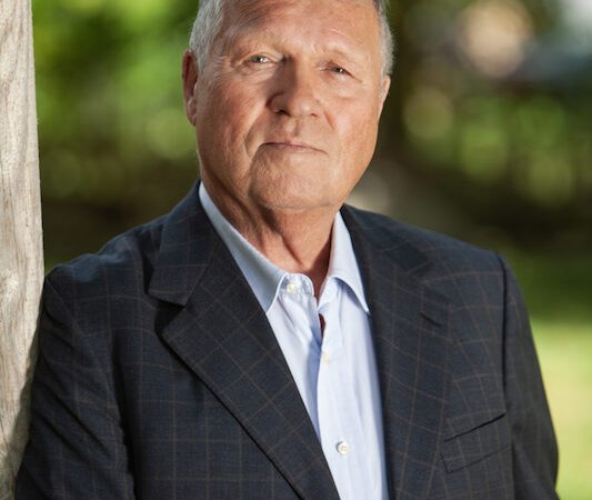 Ein &auml;lterer Mann in einem karierten Anzug lehnt an einem Baumstamm. Der Hintergrund ist unscharf und gr&uuml;n, was auf eine Au&szlig;enaufnahme hindeutet., &copy; Renitenztheater Stuttgart e.V.