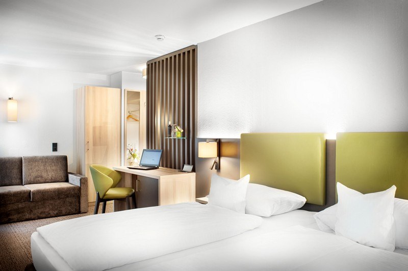 A modern hotel room with a double bed, a green headboard, a desk with a laptop, a sofa, and a wardrobe. Bright, cheerful decor., &copy; TOMAS