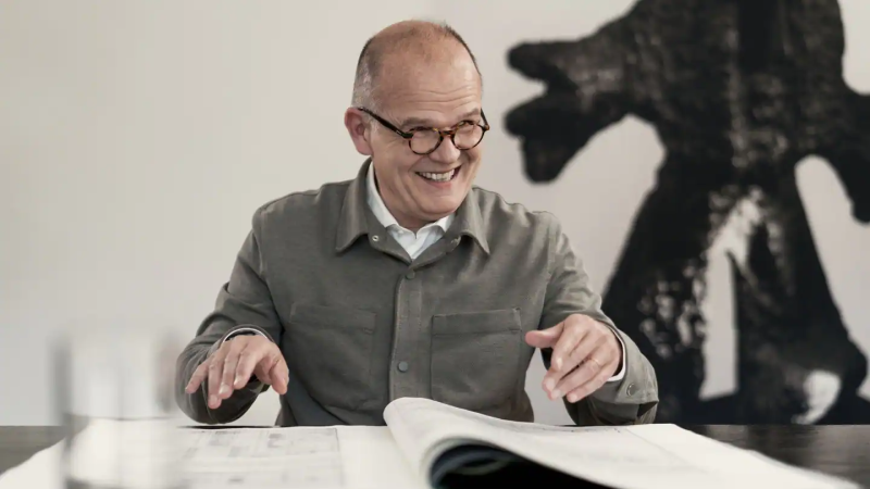 Ein Mann mit Brille l&auml;chelt und gestikuliert &uuml;ber einem aufgeschlagenen Buch. Im Hintergrund ist ein Kunstwerk zu sehen., &copy; Wolf-Peter Steinheisser