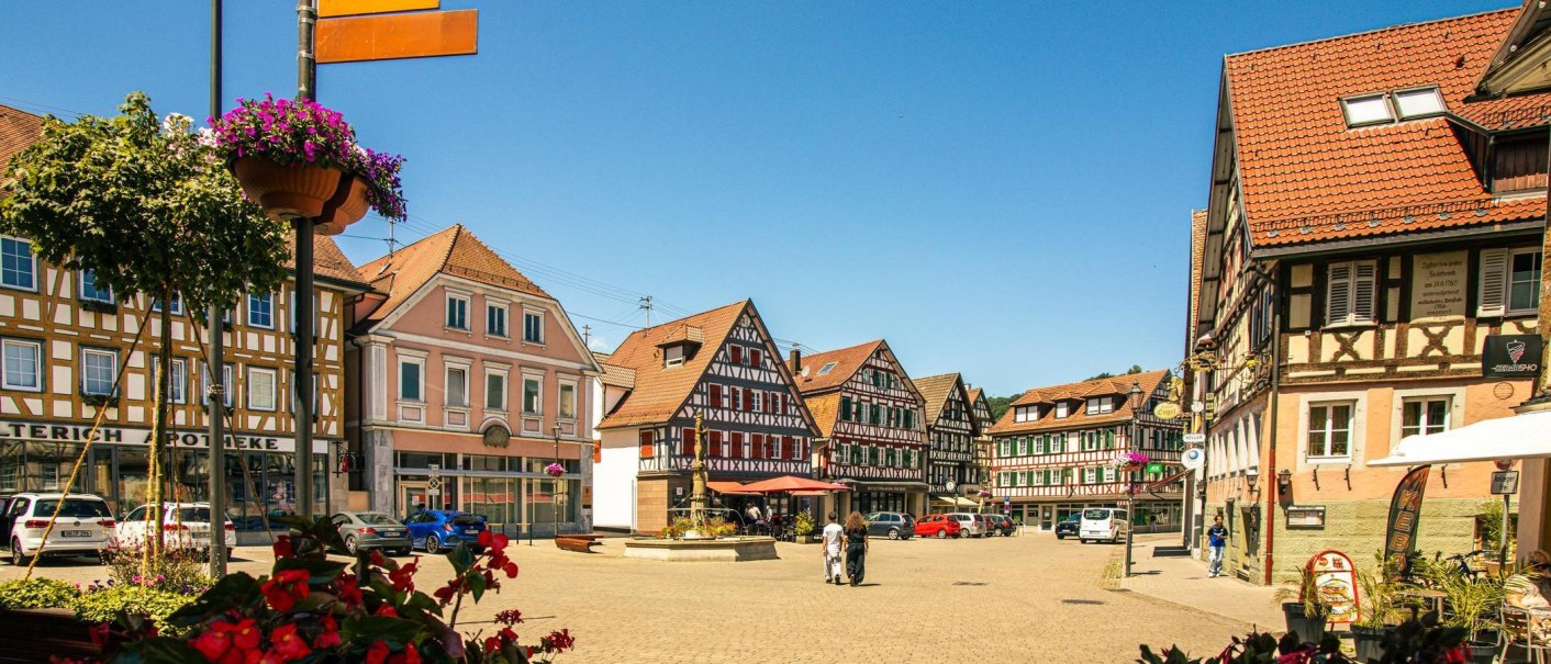 The market square in Murrhardt features picturesque half-timbered houses and a central fountain under a blue sky. Flowers adorn the square., © Stuttgart-Marketing GmbH, Sarah Schmid The market square in Murrhardt features picturesque half-timbered houses and a central fountain under a blue sky. Flowers adorn the square., © Stuttgart-Marketing GmbH, Sarah Schmid