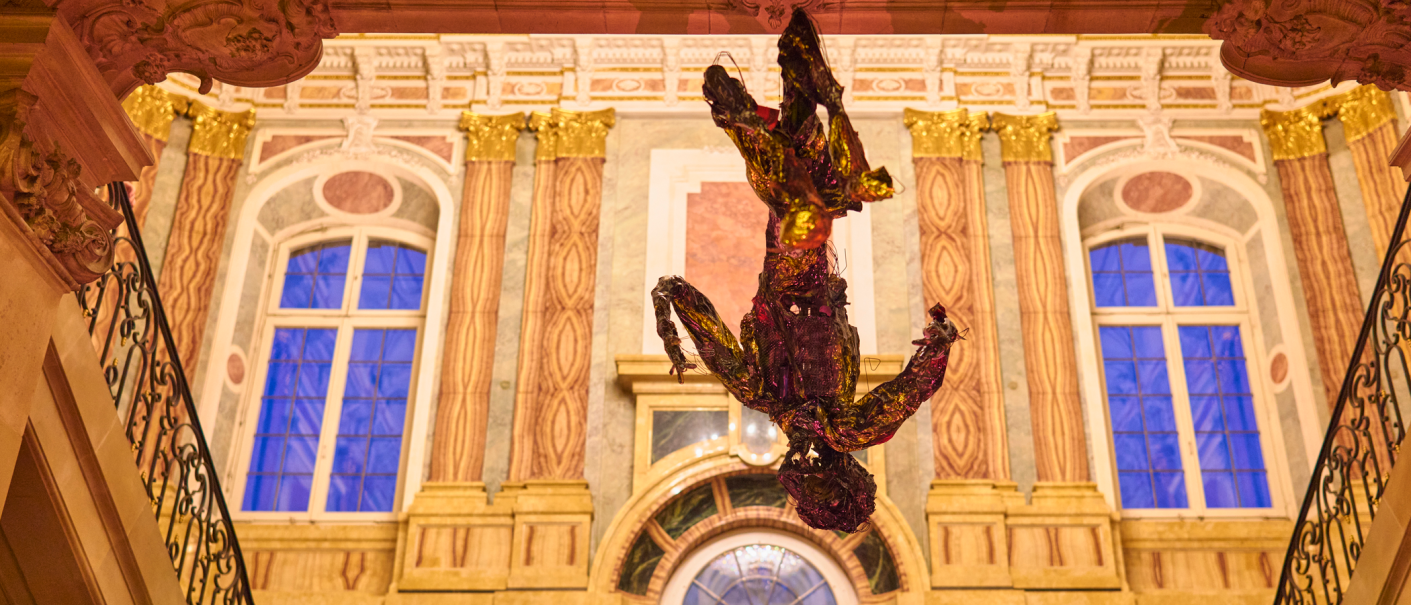 A colorful sculpture hangs upside down in a magnificent room with ornate walls and large windows. The architecture is richly decorated., &copy; PV Projekt Verlag GmbH