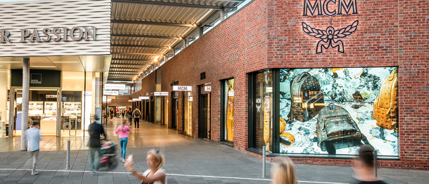 Shopping street in the Outletcity Metzingen with stores like MCM. People stroll past, some are blurred in motion., © www.johannesvogt.de Shopping street in the Outletcity Metzingen with stores like MCM. People stroll past, some are blurred in motion., © www.johannesvogt.de