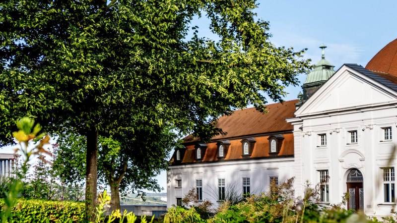 Ein historisches Gebäude mit rotem Dach und weißen Wänden, umgeben von grünen Bäumen und Pflanzen, unter einem klaren blauen Himmel., © DLA Marbach (Anja Bleeser) Ein historisches Gebäude mit rotem Dach und weißen Wänden, umgeben von grünen Bäumen und Pflanzen, unter einem klaren blauen Himmel., © DLA Marbach (Anja Bleeser)