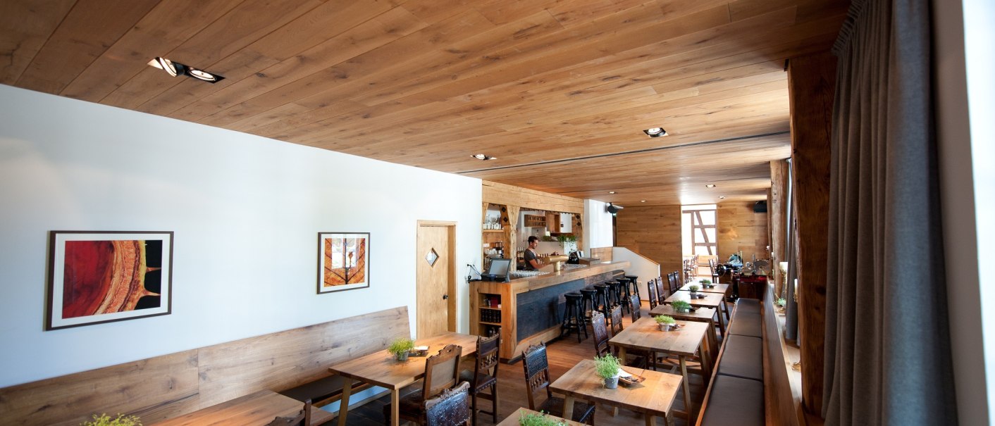Gemütliches Restaurant mit Holzmöbeln, langen Tischen und Bar. Pflanzen als Dekoration, warme Beleuchtung und Bilder an den Wänden., © Ruthenberg&Schlatter GbR Gemütliches Restaurant mit Holzmöbeln, langen Tischen und Bar. Pflanzen als Dekoration, warme Beleuchtung und Bilder an den Wänden., © Ruthenberg&Schlatter GbR