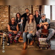 Six band members sit and stand in a rustic room. They are wearing casual clothes and smiling at the camera. Brick walls are visible in the background., © links im Bild Six band members sit and stand in a rustic room. They are wearing casual clothes and smiling at the camera. Brick walls are visible in the background., © links im Bild
