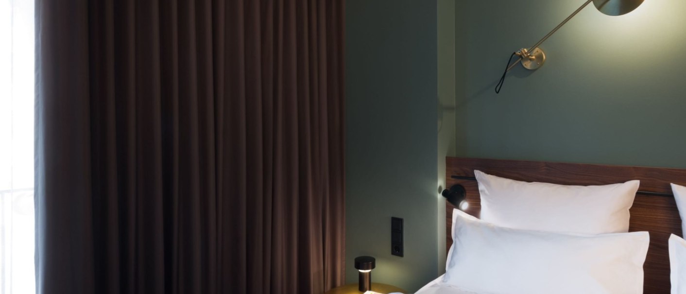 Modernes Hotelzimmer mit grüner Wand, braunen Vorhängen und stilvoller Wandlampe über dem Bett. Weiße Bettwäsche und ein kleiner Nachttisch sind sichtbar., © EmiLu Hotel GmbH