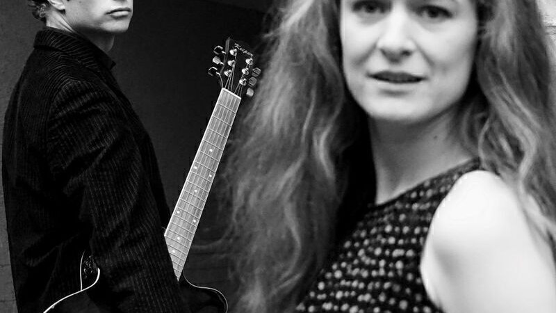 Schwarz-weiß Bild: Eine Frau im Vordergrund, unscharf, und ein Mann mit Gitarre im Hintergrund, scharf. Beide schauen in die Kamera., © Theaterhaus Stuttgart e.V.