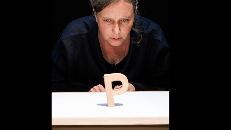 Eine Frau schaut intensiv auf einen aufrecht stehenden Buchstaben P auf einem Tisch. Der Hintergrund ist dunkel, was den Fokus auf die Szene lenkt., &copy; FITZ Das Theater animierter Formen