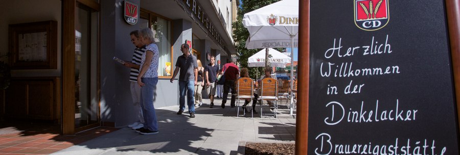 Entrance to the Dinkelacker brewery pub with a welcome sign. People walk past, some sitting at tables under parasols., © Familienbrauerei Dinkelacker