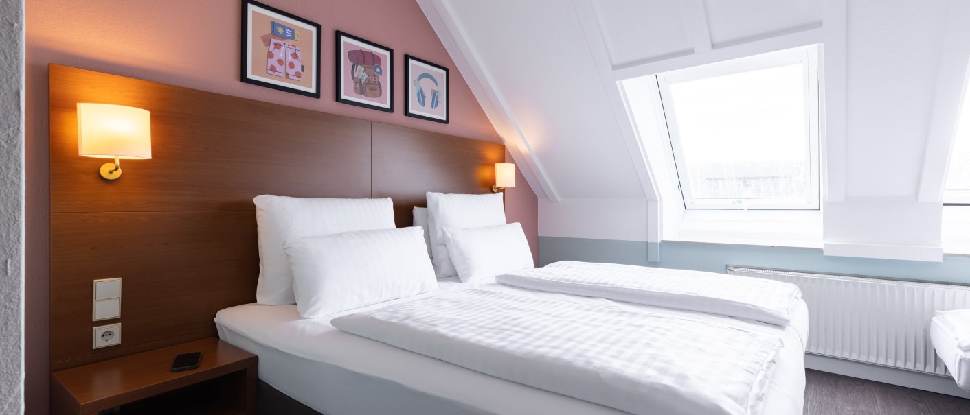 A bright bedroom with a double bed, white sheets, and a wooden headboard. Three pictures hang on the wall, and sconces are mounted next to them., &copy; Novum  Hospitality