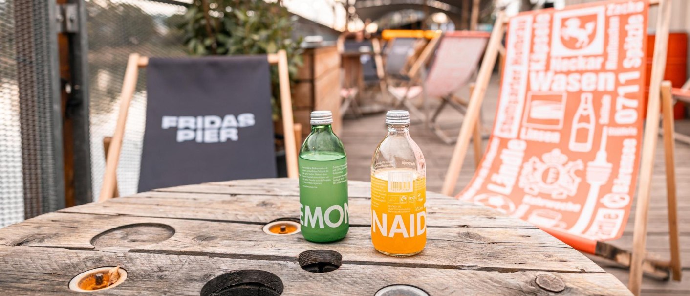 Zwei Limonadenflaschen auf einem Holztisch, umgeben von Liegestühlen bei Fridas Pier. Entspannte Atmosphäre im Freien., © SMG, Sarah Schmid
