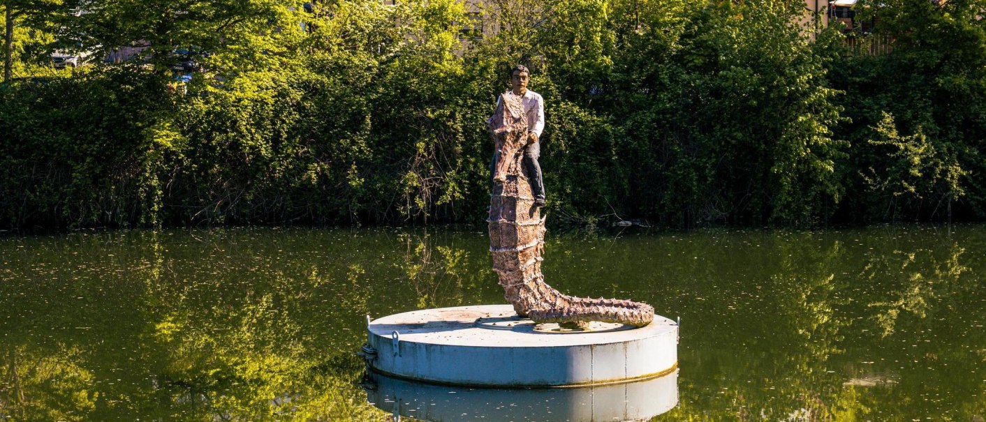 A sculpture of a man with a long tail stands on a platform in the water, surrounded by lush greenery and trees., © Stuttgart-Marketing GmbH, Sarah Schmid A sculpture of a man with a long tail stands on a platform in the water, surrounded by lush greenery and trees., © Stuttgart-Marketing GmbH, Sarah Schmid
