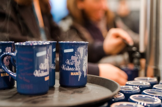 Blaue Tassen mit 'Welt Weihnachts Markt' stehen auf einem Tablett. Im Hintergrund schenken Menschen Glühwein ein, was eine festliche Atmosphäre schafft., © Cool-Tours StattReisen