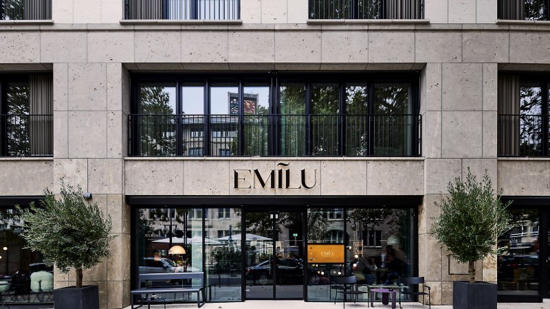 Eingang des EmiLu Hotels mit gro&szlig;en Glasfenstern, Pflanzen in schwarzen T&ouml;pfen und dem Hotelnamen &uuml;ber der T&uuml;r., &copy; EmiLu Hotel GmbH