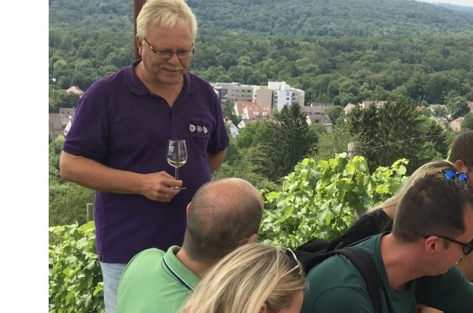 Ein Mann in lila Hemd hält ein Weinglas und spricht mit einer Gruppe von Menschen in einem Weinberg. Im Hintergrund sind Bäume und Gebäude zu sehen., © Weingenuss Stuttgart Ein Mann in lila Hemd hält ein Weinglas und spricht mit einer Gruppe von Menschen in einem Weinberg. Im Hintergrund sind Bäume und Gebäude zu sehen., © Weingenuss Stuttgart
