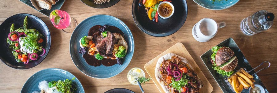 Ein Tisch mit verschiedenen Gerichten: Fleisch mit Gemüse, Salate, Burger mit Pommes, Brot und Desserts. Getränke sind ebenfalls zu sehen., © SatzDrei