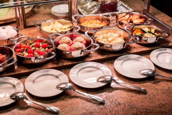Frühstücksbuffet mit Metallschalen voller Speisen, darunter Tomaten und Brötchen. Leere Teller und Löffel sind davor platziert., © Kronen Hotel GmbH Frühstücksbuffet mit Metallschalen voller Speisen, darunter Tomaten und Brötchen. Leere Teller und Löffel sind davor platziert., © Kronen Hotel GmbH