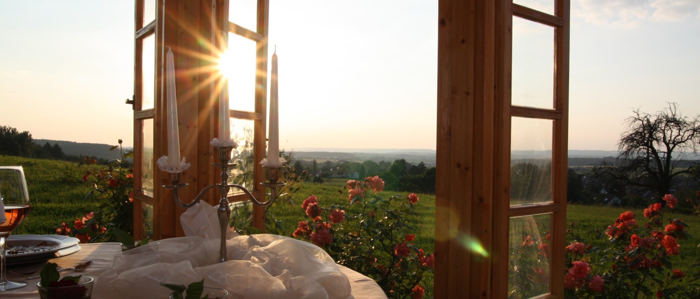 Ein gedeckter Tisch mit Kerzen und Gläsern vor einem offenen Fenster. Draußen blühende Rosen und ein Sonnenuntergang über einer grünen Wiese., © Badhotel Restaurant Stauferland
