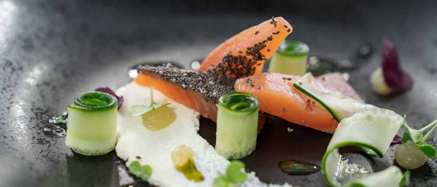 A gourmet dish featuring salmon, chia seeds, cucumber rolls, and cream, artfully arranged on a black plate., &copy; Flairhotel Vier Jahreszeiten