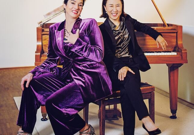 Zwei Frauen in eleganter Kleidung sitzen l&auml;chelnd vor einem Klavier. Eine tr&auml;gt einen lila Anzug, die andere einen schwarzen Anzug., &copy; Wilhelma Theater