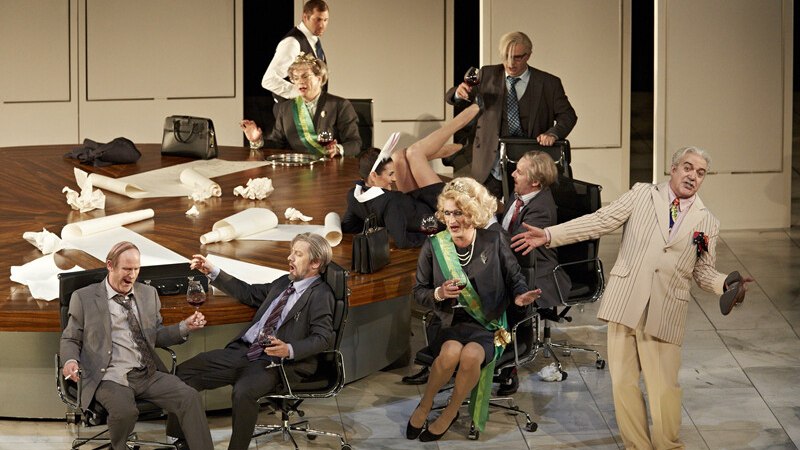 Opernszene mit Darstellern in Anzügen und Kostümen um einen Tisch. Einige sitzen, andere stehen, alle wirken in eine lebhafte Diskussion vertieft., © Württembergische Staatstheater Stuttgart Opernszene mit Darstellern in Anzügen und Kostümen um einen Tisch. Einige sitzen, andere stehen, alle wirken in eine lebhafte Diskussion vertieft., © Württembergische Staatstheater Stuttgart