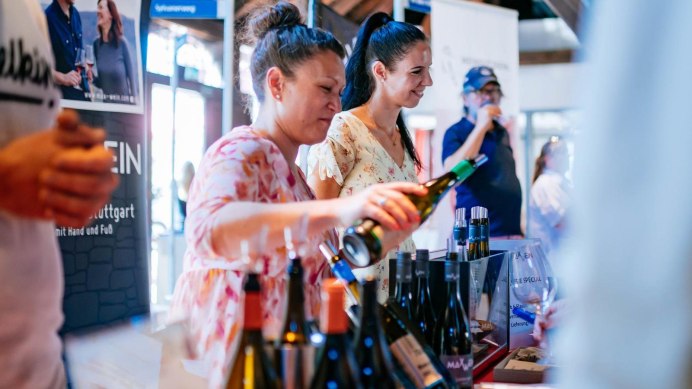 Two women serve wine at a tasting event, surrounded by wine bottles. In the background there are some guests tasting wine at a different table., © Stuttgart-Marketing GmbH, Thomas Niedermüller