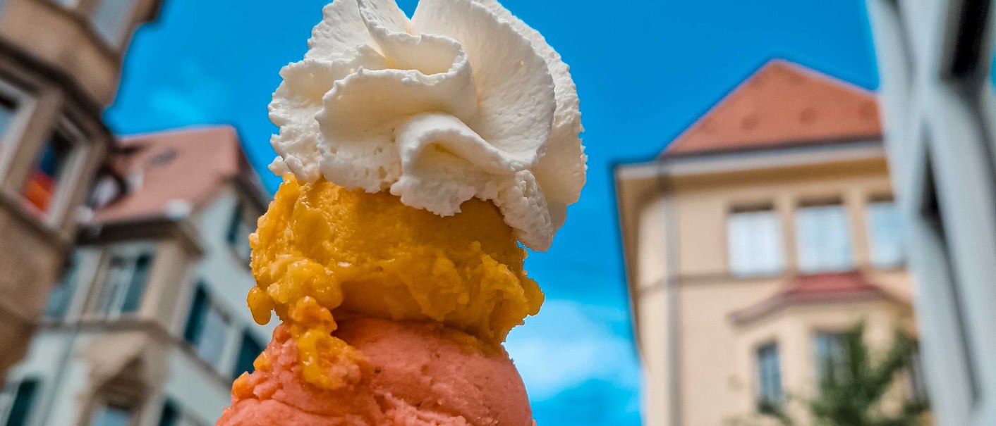 Eine Eist&uuml;te mit drei Kugeln und Sahne vor einem blauen Himmel und unscharfen Geb&auml;uden im Hintergrund., &copy; Kesselgl&uuml;ck