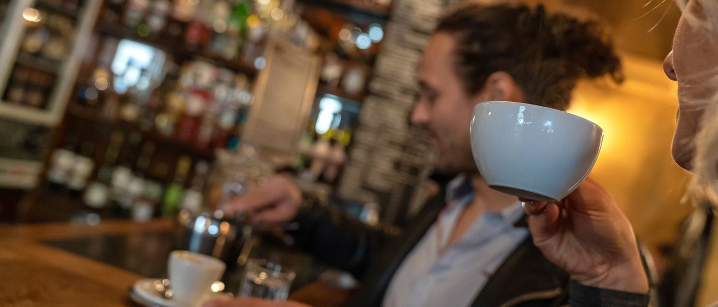 Two people in a café. One person is drinking from a cup while the other prepares coffee. Shelves with bottles can be seen in the background., © Stuttgart Marketing  GmbH, Fotografin Martina Denker, post@denkerfotografie.de
