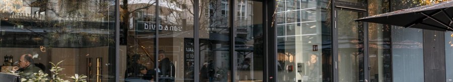 Eingang eines Cafés mit Glasfront und Außenbestuhlung. Pflanzen hängen von oben herab. Menschen sitzen drinnen. Schild mit 'sotto sopra' sichtbar., © SMG, Sarah Schmid