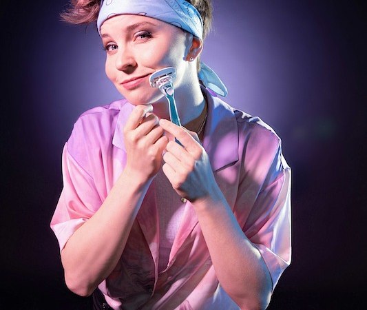 Person mit Bandana und Rasierer posiert l&auml;chelnd vor einem lila Hintergrund. Sie tr&auml;gt ein rosa Hemd und hat kurzes, hochgestyltes Haar., &copy; Rosenau Kultur e.V.