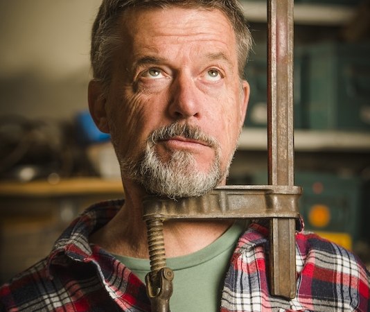 Ein Mann mit grauem Bart und kariertem Hemd hält eine große Schraubzwinge um seinen Kopf. Er blickt nach oben, im Hintergrund sind Werkzeuge zu sehen., © Rosenau Kultur e.V. Ein Mann mit grauem Bart und kariertem Hemd hält eine große Schraubzwinge um seinen Kopf. Er blickt nach oben, im Hintergrund sind Werkzeuge zu sehen., © Rosenau Kultur e.V.