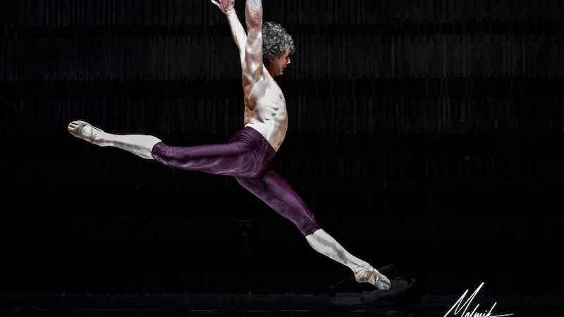 Ein Balletttänzer in violetter Kleidung führt einen beeindruckenden Sprung auf einer dunklen Bühne aus., © István Simon