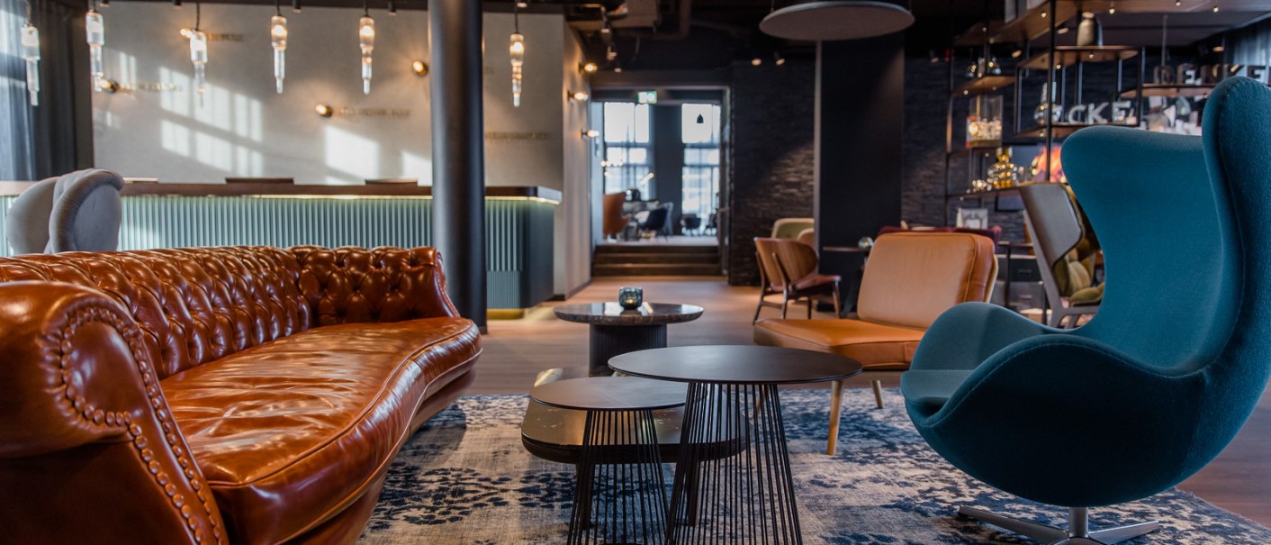 Moderne Hotellobby mit braunem Ledersofa, blauem Sessel, Couchtischen und stilvoller Beleuchtung. Elegantes, gemütliches Ambiente., © TOMAS