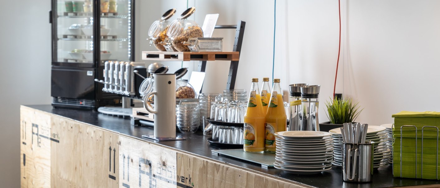 Ein Frühstücksbereich mit Getränken, Geschirr und Snacks auf einem Holztresen. Im Hintergrund ein Kühlschrank mit weiteren Speisen., © a2 HOTELS Ein Frühstücksbereich mit Getränken, Geschirr und Snacks auf einem Holztresen. Im Hintergrund ein Kühlschrank mit weiteren Speisen., © a2 HOTELS