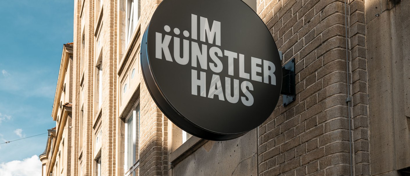 Rundes Schild mit 'IM KÜNSTLER HAUS' an einer Backsteinfassade, blauer Himmel im Hintergrund., © SMG, Sarah Schmid