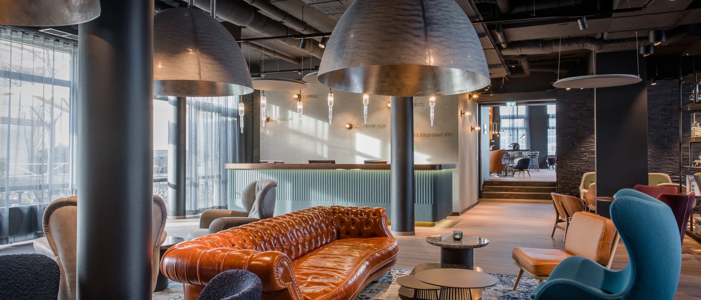 Elegante Hotellobby mit braunem Ledersofa, blauen und grauen Sesseln, großen Hängelampen und einem Empfangstresen im Hintergrund., © TOMAS