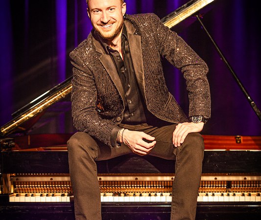 Ein Mann in glitzerndem Sakko sitzt l&auml;chelnd auf einem Klavier vor lila Vorh&auml;ngen., &copy; Renitenztheater Stuttgart e.V.