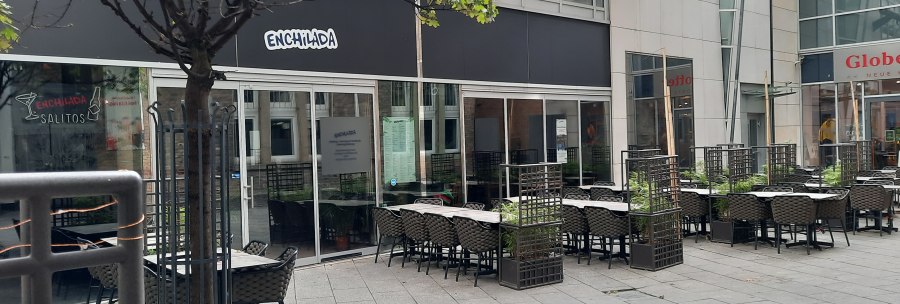 Outdoor area of a restaurant called Enchilada with empty tables and chairs. A tree and plants separate the seating areas., &copy; SMG
