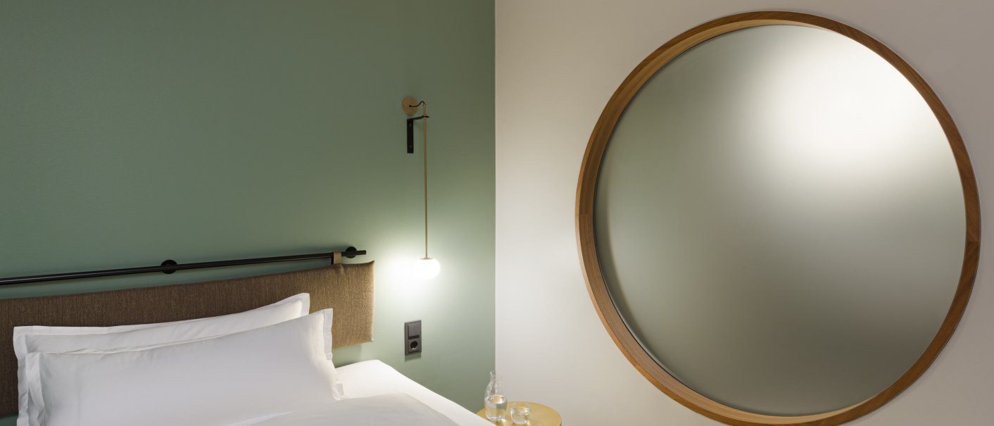 Ein Bett mit weißer Bettwäsche, eine runde Wandlampe und ein großer runder Spiegel an einer grünen Wand., © EmiLu Hotel GmbH